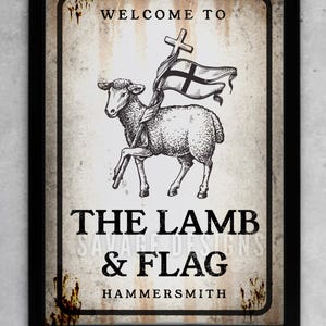 May include: A framed sign with a vintage aesthetic. The sign features a lamb holding a flag with a cross, and the text "WELCOME TO THE LAMB & FLAG HAMMERSMITH". The design includes the text "SAVAGE DESIGNS".