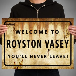 Puede incluir: Un cartel de metal oxidado con el texto "Bienvenido a Royston Vasey ¡Nunca te irás!" El cartel lo sostiene una persona que lleva una sudadera con capucha negra.