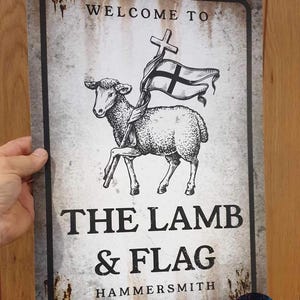 May include: A rectangular sign with a distressed, weathered appearance. The sign features the text "WELCOME TO" at the top, and below, a detailed illustration of a lamb holding a flag with a cross. The text "THE LAMB & FLAG" is prominently displayed.