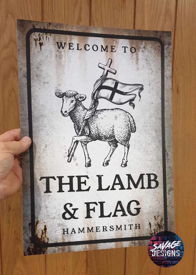 May include: A rectangular sign with a distressed, weathered appearance. The sign features the text "WELCOME TO" at the top, and below, a detailed illustration of a lamb holding a flag with a cross. The text "THE LAMB & FLAG" is prominently displayed.