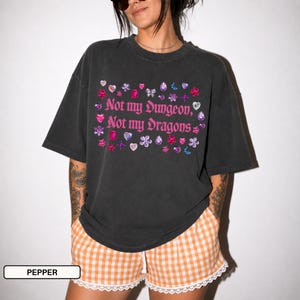 Könnte beinhalten: Ein anthrazitfarbenes Kurzarm-T-Shirt mit dem rosa Text "Not my Dungeon, Not my Dragons", umgeben von einem verstreuten Muster aus bunten Edelstein-, Herz-, Blumen- und Sternmotiven. Das T-Shirt hat einen Rundhalsausschnitt und eine lockere Passform.