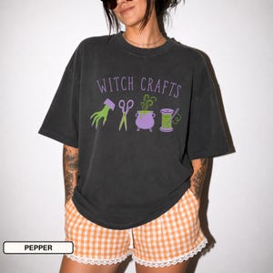 Può includere: Una t-shirt grigio antracite a maniche corte con la scritta WITCH CRAFTS in lettere viola sopra un'immagine grafica di una mano verde, forbici, un calderone viola con vapore verde e un rocchetto di filo verde con un ago.