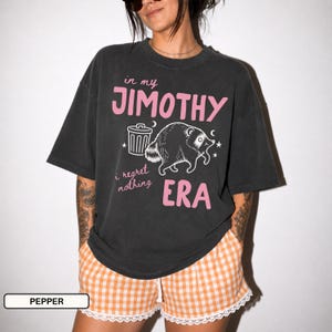Può includere: Una t-shirt oversize grigio antracite con la grafica di un procione accanto a un bidone della spazzatura. Il testo recita in my JIMOTHY ERA e i regret nothing in lettere rosa. La maglietta è etichettata come colore pepper, ha maniche corte e girocollo.