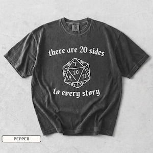 May include: A charcoal grey short-sleeve t-shirt featuring a white graphic of a twenty-sided die in the centre. The text "there are 20 sides" is printed above the die, and "to every story" is printed below the die in a white serif font.