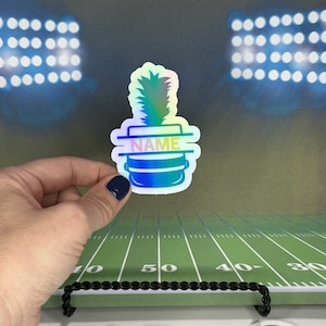 Puede incluir: Una pegatina holográfica de un sombrero de banda de marcha con una pluma y la palabra "NAME" en una pancarta. La pegatina está en un campo de fútbol verde y blanco.