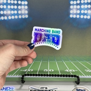 May include: A holographic sticker with the text "Marching Band Dad" in blue and green with a musical note and a trumpet. The sticker is on a green and white football field.