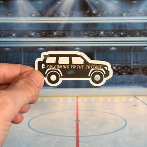 May include: A black and white sticker in the shape of a car with the text "I'M COMING TO THE COTTAGE." The sticker has a white border and is held in front of a hockey rink background.