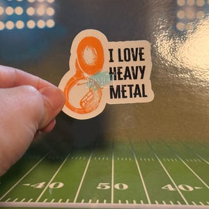 May include: An orange and white sticker featuring the text "I LOVE HEAVY METAL" and a sousaphone illustration. The sticker is held by a hand against a backdrop of a football pitch.
