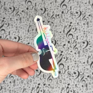 May include: A holographic sticker in the shape of a double bass, with the name "Ashlyn" written in cursive. The sticker has a rainbow gradient effect and is held against a background of musical notes.
