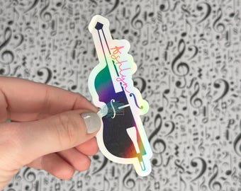 May include: A holographic sticker in the shape of a double bass, with the name "Ashlyn" written in cursive. The sticker has a rainbow gradient effect and is held against a background of musical notes.