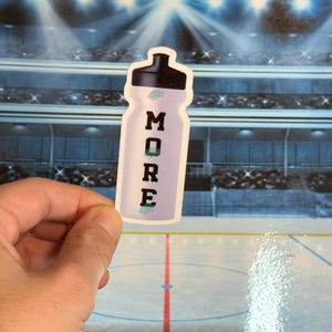 May include: A sticker of a water bottle with the word "MORE" in black capital letters. The bottle is light purple with a black cap. The background is a blurred image of a hockey arena.