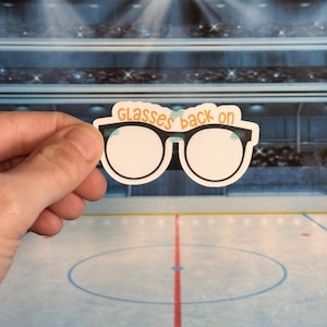 May include: A sticker featuring a pair of eyeglasses with the text "Glasses back on" in orange. The glasses have a black frame and white lenses. The sticker is held against a background of an ice hockey rink.