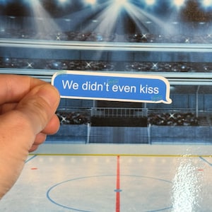 Peut inclure: Un autocollant bleu en forme de bulle de message texte avec les mots "We didn't even kiss" en texte blanc. L'autocollant est tenu devant un fond représentant une patinoire de hockey sur glace.