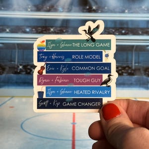 May include: A sticker with text and illustrations. The sticker has multiple rectangular sections with names and phrases like "THE LONG GAME" and "ROLE MODEL." Illustrations of a violin, a drink, and birds are included. The sticker is held by a hand.