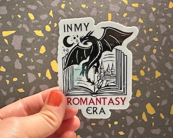 May include: A sticker with a black dragon flying out of an open book. The book has a castle in the background. The text on the sticker reads "In My Romantasy Era".