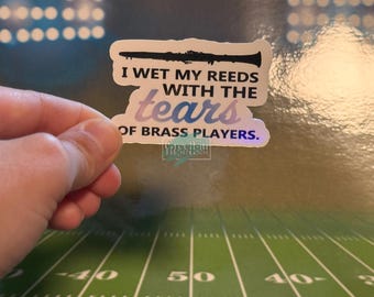 May include: A white sticker with the text "I WET MY REEDS WITH THE tears OF BRASS PLAYERS." features a black clarinet illustration. The word "tears" is in a gradient of blue and purple. The sticker is held by a hand.