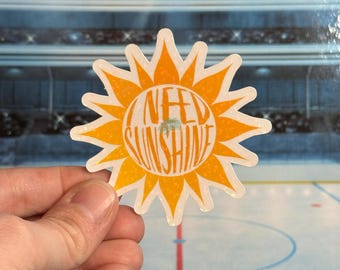 Könnte beinhalten: Ein gestanzter Aufkleber in Form einer Sonne mit orangefarbenen Strahlen und einem gelben Zentrum. Der Text "I NEED SUNSHINE" ist orangefarben im Inneren der Sonne aufgedruckt. Der Aufkleber wird von einer Hand gehalten.