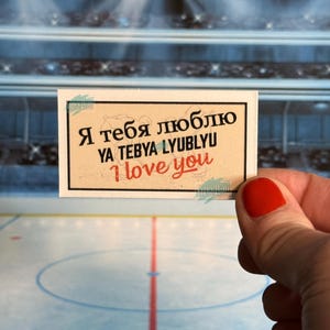 May include: A rectangular sticker with a tan background and black border. The sticker has text in Russian and English that says "I love you". The sticker is held in front of a hockey rink.