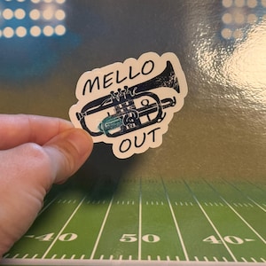 May include: A sticker with a black and teal illustration of a trumpet with the words "MELLO OUT" in a bold font. The sticker is held by a hand against a background that resembles a football field.