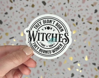 Puede incluir: Una pegatina redonda blanca con texto negro: "THEY DIDN'T BURN WITCHES THEY BURNED WOMEN". La pegatina tiene un borde negro y elementos decorativos. El fondo presenta un patrón moteado.