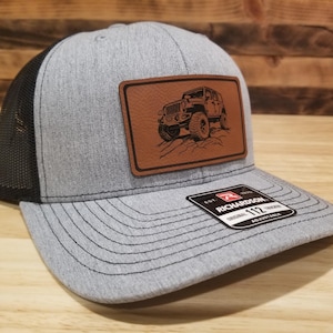 May include: Gray and black trucker hat with a brown leather patch featuring an off-road vehicle illustration. The hat has a curved brim and an adjustable snap closure. The brand tag is visible.
