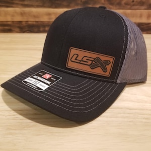 May include: Black and gray trucker hat with a leather patch featuring the letters "LSX" in a brown color. The hat has a curved brim and a mesh back. The Richardson 112 Trucker hat is adjustable.