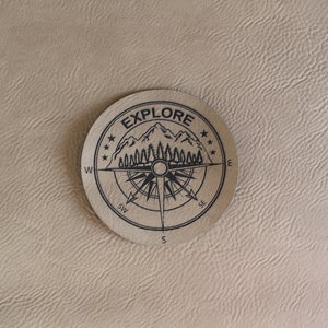 May include: A round, tan leather patch with the word "EXPLORE" in a circular design. The design includes a compass rose, mountains, trees, and stars, all in black. The patch is set against a textured, beige background.