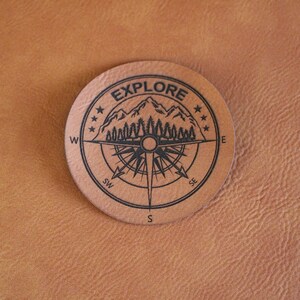 May include: A round, tan leather coaster with a compass design and the word "EXPLORE" in a circular border. The compass features a mountain range and trees. The background is a textured, tan surface.