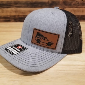 May include: Gray trucker hat with a black mesh back and a brown leather patch featuring a vehicle graphic. The hat has a curved brim and an adjustable snap closure. The brand name "Richardson" is visible on a tag.
