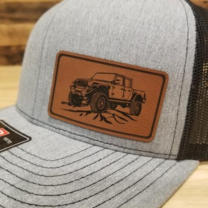 May include: Gray and black trucker hat with a brown leather patch featuring a detailed illustration of a truck. The hat has a curved brim and a mesh back. The patch has a stitched border.