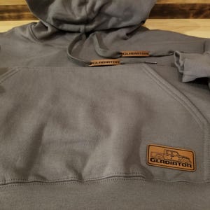 May include: A gray hoodie with a drawstring hood and a kangaroo pocket. The hoodie features a brown leather patch with a truck graphic and the word "GLADIATOR". The drawstring ends also have "GLADIATOR" printed on them.
