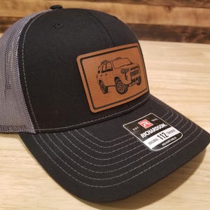 May include: Black trucker hat with a gray mesh back and a brown leather patch featuring a vehicle illustration. The hat has a curved brim and an adjustable strap. The Richardson 112 hat is a classic style.