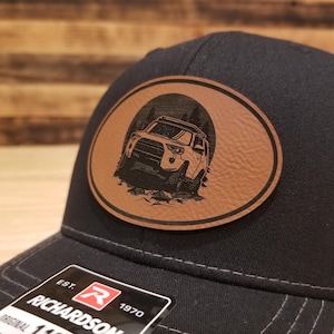 May include: Black trucker hat with a brown leather patch featuring an off-road vehicle graphic. The patch is oval-shaped with a detailed illustration of a 4x4 vehicle in a natural setting. The hat includes a Richardson tag.
