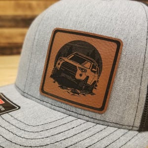 May include: Gray trucker hat with a black mesh back and a square, brown leather patch. The patch features a black illustration of an off-road vehicle in front of a forest and a sun. The hat has a Richardson 1970 tag.