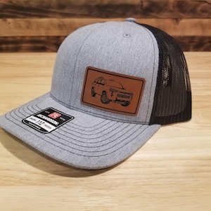 May include: Gray trucker hat with a black mesh back and a brown leather patch featuring an engraved off-road vehicle. The hat has a curved brim and a Richardson 112 tag.