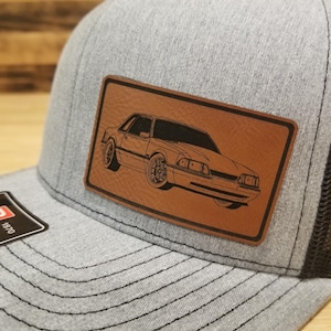 May include: A gray trucker hat with a black mesh back and a brown leather patch. The patch features a black outline drawing of a classic car. The hat has a curved brim and a Richardson tag.