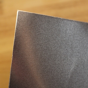 May include: Close-up of a textured, dark gray rectangular object with a metallic sheen. The edge of the object is visible, showing a clean, sharp cut. The background is a blurred wooden surface.