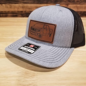 May include: Gray and black trucker hat with a brown leather patch featuring a truck outline. The hat has a curved brim and a mesh back. The Richardson 112 adjustable hat is displayed on a wooden surface.