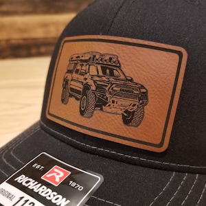 May include: Black trucker hat with a brown leather patch featuring a detailed illustration of an off-road truck. The hat has a curved brim and a Richardson 112 tag.