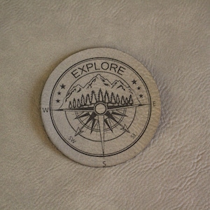 May include: A round, light gray coaster with the word "EXPLORE" in a circular border. The design includes a compass rose, mountains, trees, and stars. The coaster is made of a textured material.