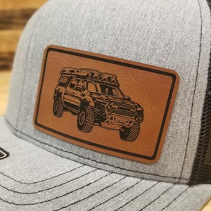 May include: A gray trucker hat with a black mesh back and a brown leather patch. The patch features a detailed black illustration of a truck with a roof rack. The hat has a curved brim and a snapback closure.