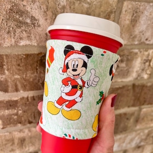 May include: A red travel mug with a white lid and a festive fabric sleeve. The sleeve features a cartoon Mickey Mouse dressed as Santa Claus, with holly and stocking accents on a light green background.