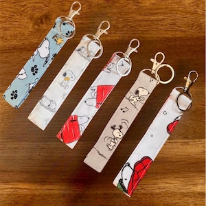 May include: Five fabric keychains with metal clasps. Each keychain features a different Snoopy design on a light blue, white, or tan background. Designs include Snoopy with paw prints, Snoopy with Woodstock, and Snoopy by his doghouse.