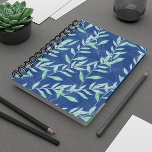 Peut inclure: Cahier à spirales avec une couverture bleue ornée d'un motif de feuilles vert clair. Le cahier est ouvert, révélant des pages vierges. Deux crayons noirs et une petite plante succulente sont également visibles.