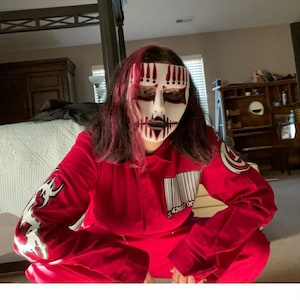 May include: A red jumpsuit featuring a white barcode graphic on the chest with the number 42617 00, a white tribal design on the right sleeve, and a circular logo on the left shoulder. The outfit is paired with a white mask with red and black markings.