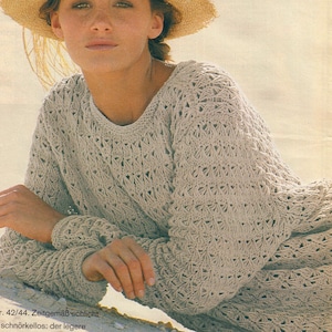 May include: A light beige, open-weave knit sweater with a crew neck and long sleeves. The sweater has a textured, patterned design. The person in the image is also wearing a straw hat.