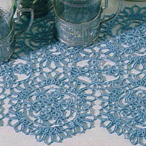 May include: A light blue, intricately crocheted table runner with a repeating floral pattern. The runner is placed on a white surface, and two silver-colored metal cup holders are visible in the background.
