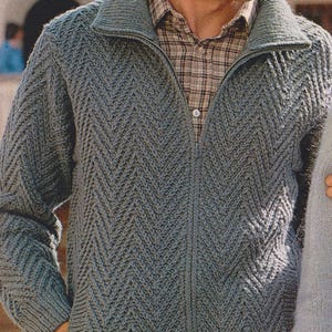 Puede incluir: Un suéter gris tejido con cremallera y un patrón de chevrón. El suéter tiene un cuello alto y bolsillos. La persona lleva una camisa a cuadros debajo. El suéter está hecho de un material suave y cómodo.