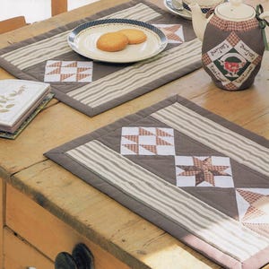 Könnte beinhalten: Zwei braune, gesteppte Tischsets mit Streifenmuster und Patchwork-Quadraten. Ein Teller mit zwei Keksen und eine Teekanne mit einer Teewärmer auf dem Holztisch. Die Tischsets haben einen braunen Rand.