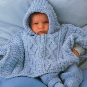 May include: A baby in a light blue knitted hooded poncho and matching trousers. The poncho has a cable knit design on the front and hood. The baby is sitting on a light blue surface.
